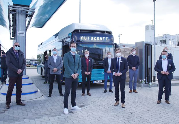 Mehrere Herren stehen vor einem Wasserstoffbus der Autokraft und einer Wasserstofftankstelle. Der Bus ist eFarm-Petrol und die Herren tragen Jackett und Krawatte.