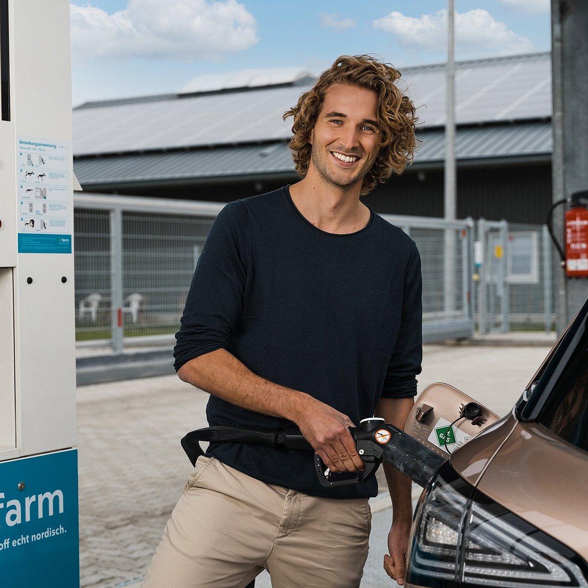 Ein junger Mann (Tim Schmitz) sateht an einer eFarm Wasserstofftankstelle und hält den Zapfrüssel in seiner Hand. Mit dem Zapfrüssel tankt er ein rosé farbenes Wasserstoffauto.